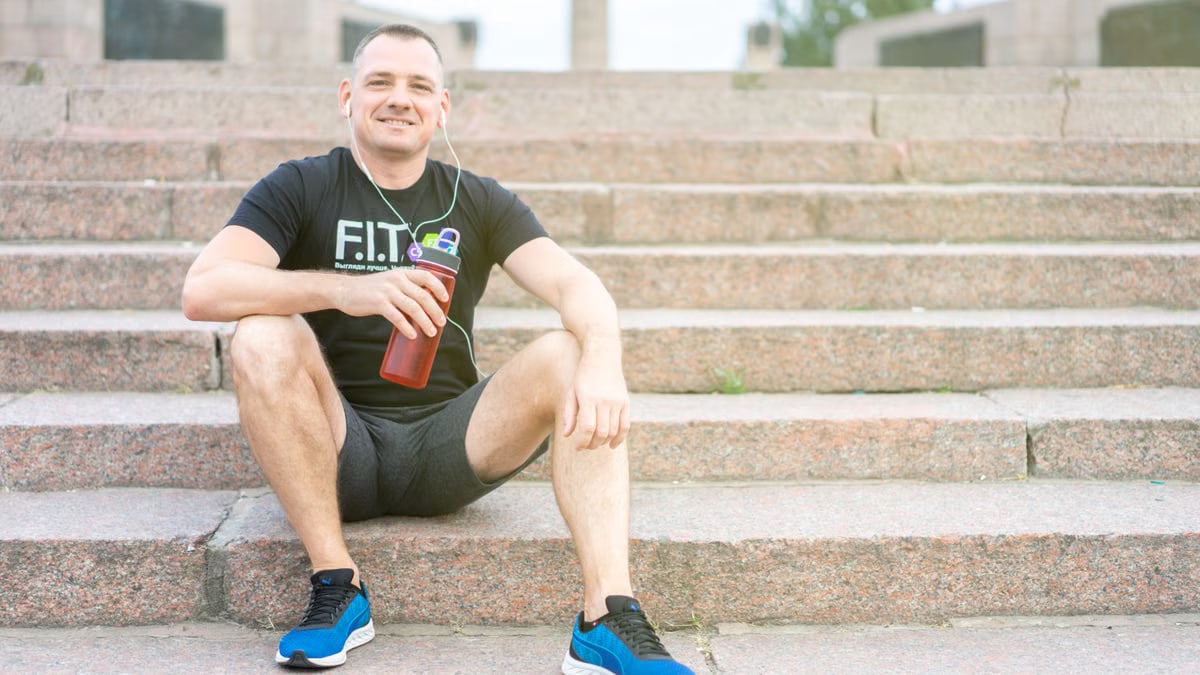 Smiling man in activewear sitting on stairs with a tumbler, enjoying a sunny day.